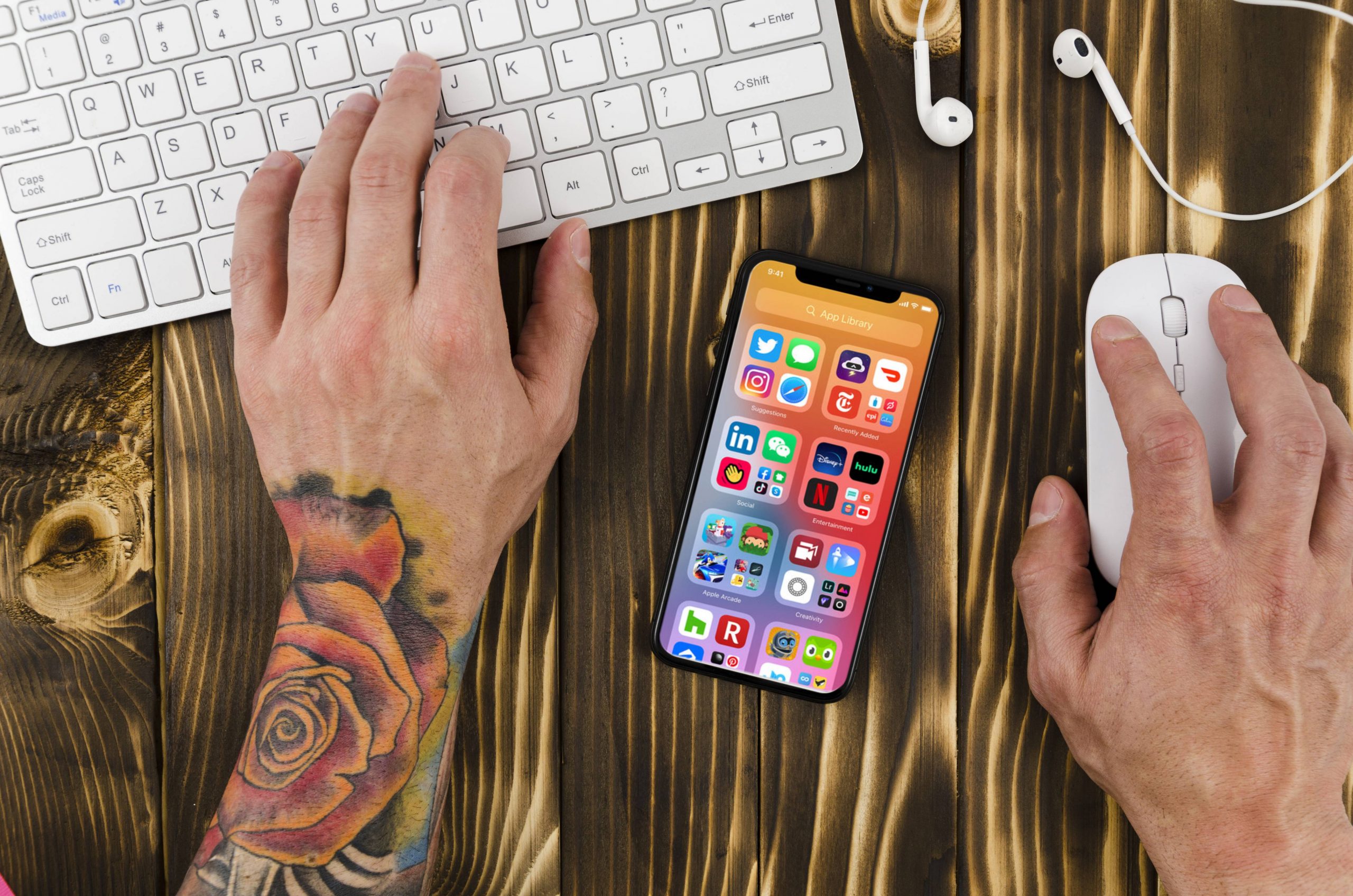 Mãos de homem tatuado mexendo num teclado de mac com um iPhone 11 cheio de apps do iOS 14